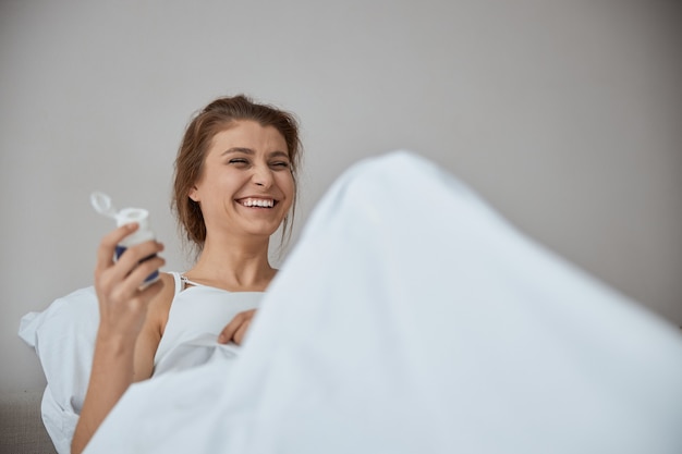 Young cheerful woman sitting in bed and leaning on pillow while going to take vitamin