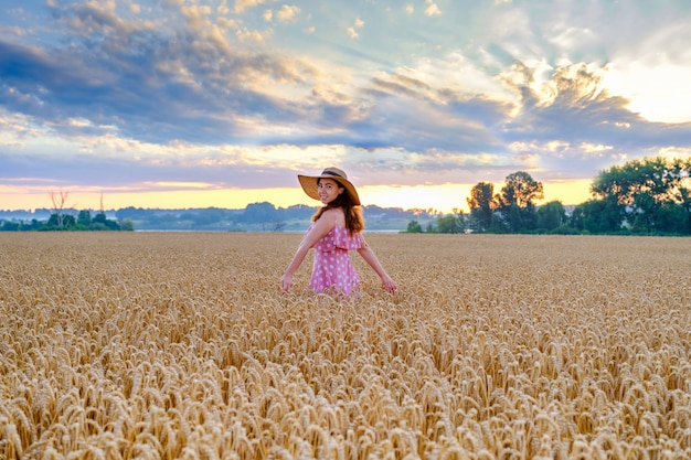 Young cheerful woman in straw hat in the middle of wheat field with outstretched arms turn around to camera and smiling.