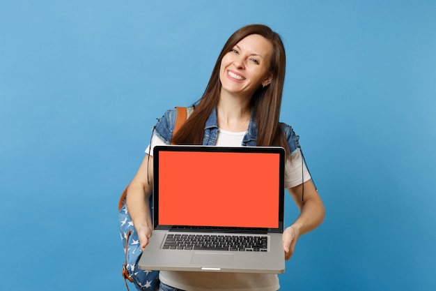 파란색 배경에 격리된 빈 검은색 빈 화면이 있는 노트북 컴퓨터를 들고 배낭을 메고 데님 옷을 입은 젊고 쾌활한 여학생입니다. 대학에서 교육입니다. 광고 공간을 복사합니다.