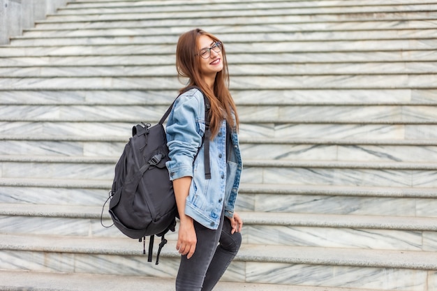 Молодая жизнерадостная студентка в джинсовой куртке и очках с большим рюкзаком поднимается по лестнице в городе. Молодежная концепция