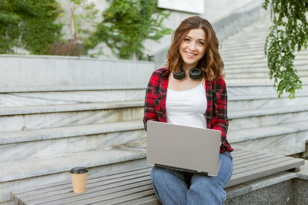 屋外でラップトップを使用して若い陽気な女性の学生。遠隔作業または教育の概念