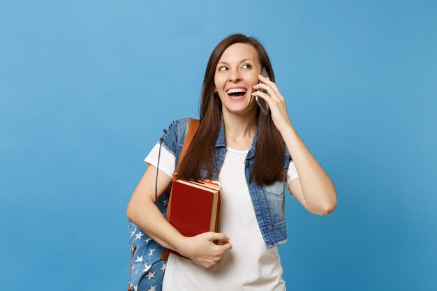バックパックを持った若い陽気な女性の学生は、青い背景で隔離の楽しい会話を行う携帯電話で話している教科書を保持しています。高校大学カレッジコンセプトの教育。