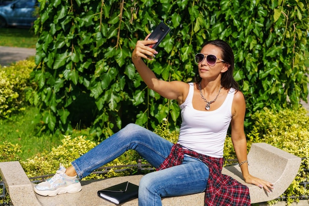 Young cheerful woman takes selfie from hands with phone while sitting on park