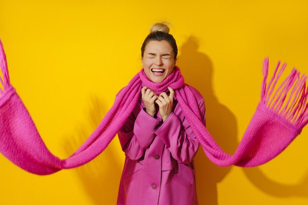 Photo young cheerful woman wearing pink coat and wool scarf against yellow background