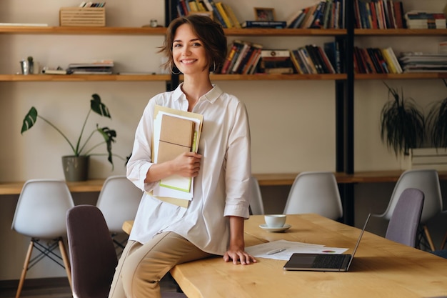 現代のオフィスで紙とラップトップと机の上に座っている間、カメラを喜んで見ている白いシャツを着た若い陽気な女性