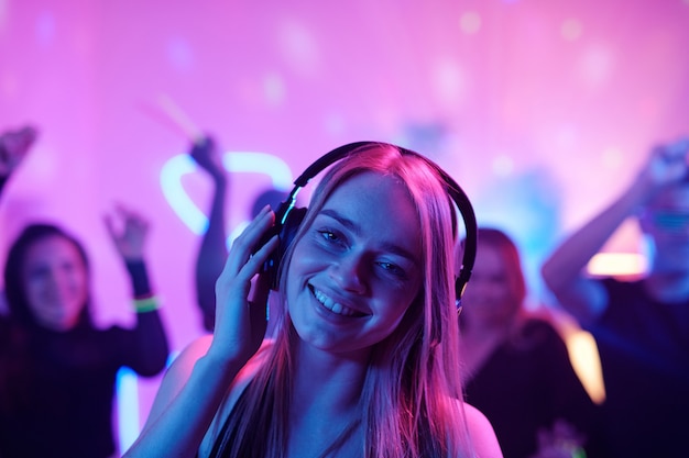 Young cheerful woman with long blond hair snapping fingers while enjoying music in headphones in front of dancing crowd at party