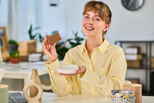 Young cheerful woman with toothy smile showing bar of soap to subscribers of her channel during livestream about self care