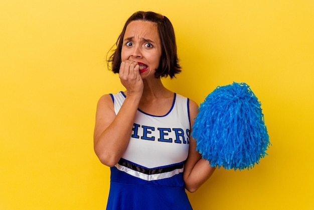 Young cheerleader mixed race woman isolated on yellow background biting fingernails, nervous and very anxious.