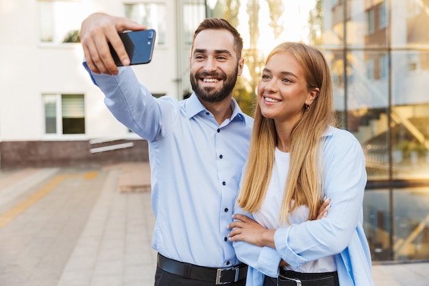 ビジネスセンターの近くの通りの外の若い陽気な幸せな男性と女性のビジネスマンは、携帯電話で自分撮りをします。