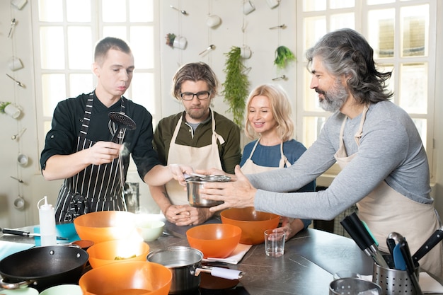 Young chef or coach in apron holding cooking tools and pan with water over table while senior man helping him among other learners