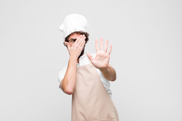 Foto giovane chef che copre il viso con la mano e mettendo l'altra mano in alto per fermare la fotocamera, rifiutando foto o immagini sul muro bianco