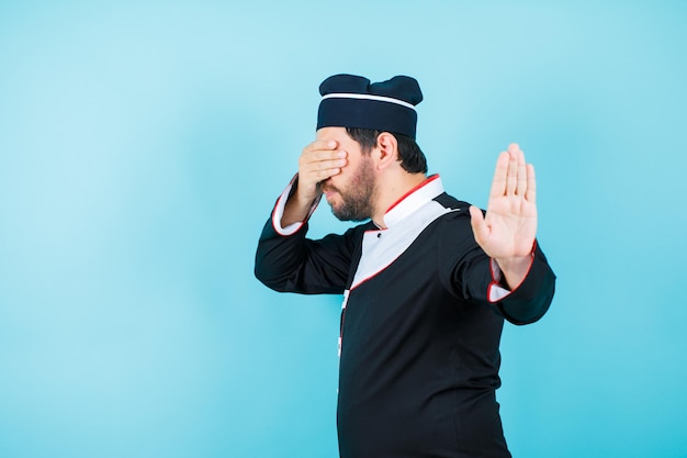 Young chef is covering his eyes and showing stop getsure with other hand on blue background