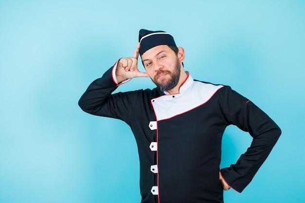 Young chef is posing to camera by holding hand on temple and putting other hand on waist on blue background