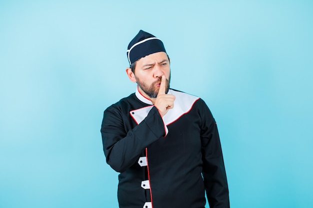 Young chef is showing silence gesture by holding fingers on lips on blue background