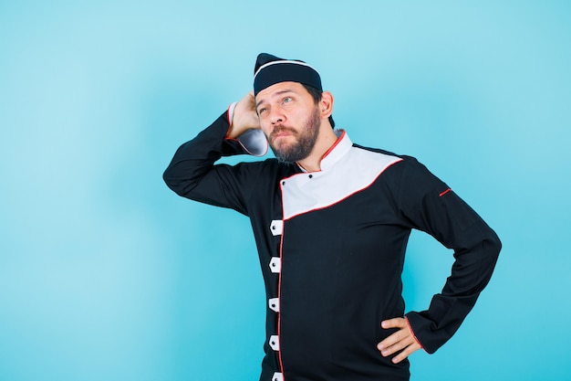 Young chef is thinking by putting hand behind head and putting other hand on waist on blue background