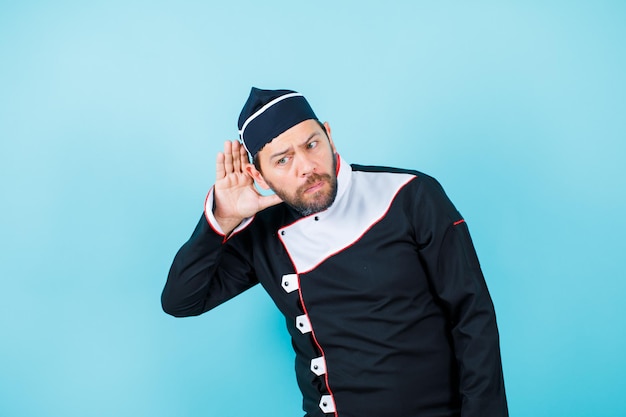 Young chef is trying to hear by holding hand near ear on blue background