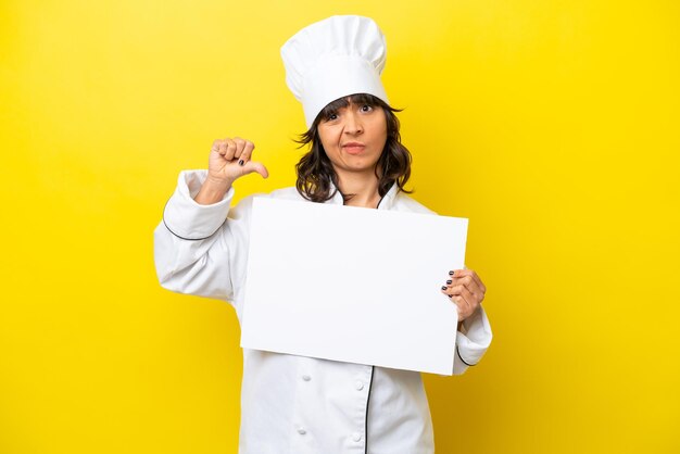 Young chef latin woman isolated on yellow background holding an empty placard and doing bad signal