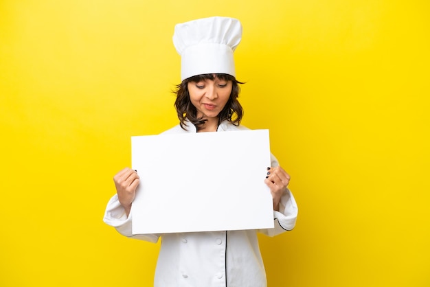 Young chef latin woman isolated on yellow background holding an empty placard and looking it