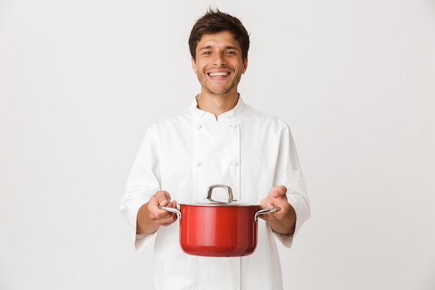 Young chef man standing isolated on white wall holding crockery.