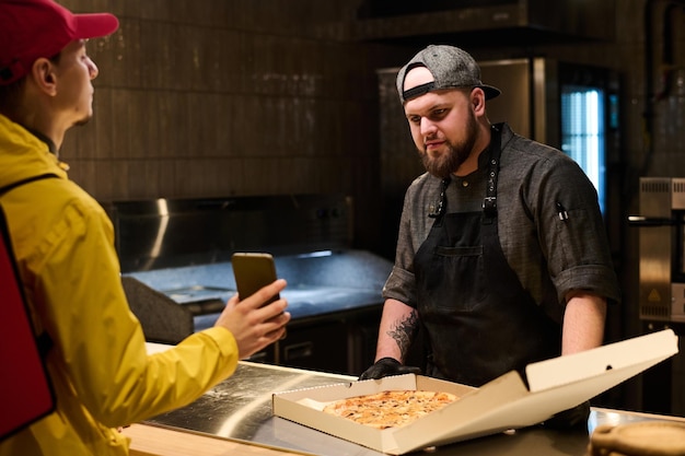 Young chef in uniform looking at smartphone screen held by deliveryman