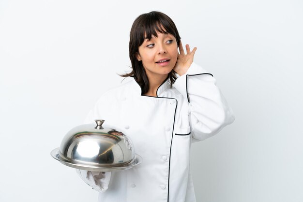 Young chef with tray isolated on white background listening to something by putting hand on the ear