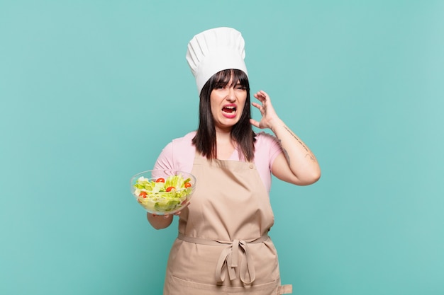 Young chef woman screaming with hands up in the air, feeling furious, frustrated, stressed and upset