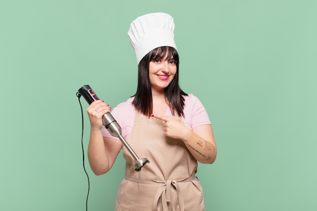 Young chef woman smiling cheerfully, feeling happy and pointing to the side and upwards, showing object in copy space