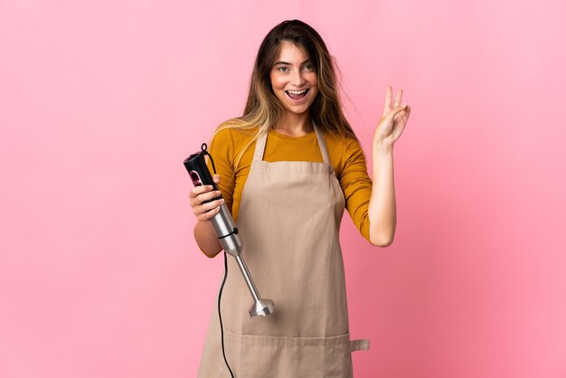 Young chef woman using hand blender isolated on pink wall smiling and showing victory sign