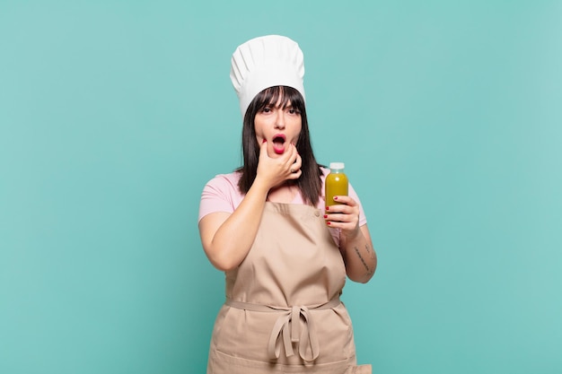 Young chef woman with mouth and eyes wide open and hand on chin, feeling unpleasantly shocked, saying what or wow