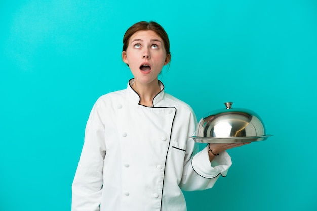 Young chef woman with tray isolated on blue background looking up and with surprised expression