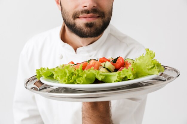 Giovane capo uomo in uniforme da cuoco piatto profumato mentre si tiene piatto con insalata di verdure isolato su muro bianco