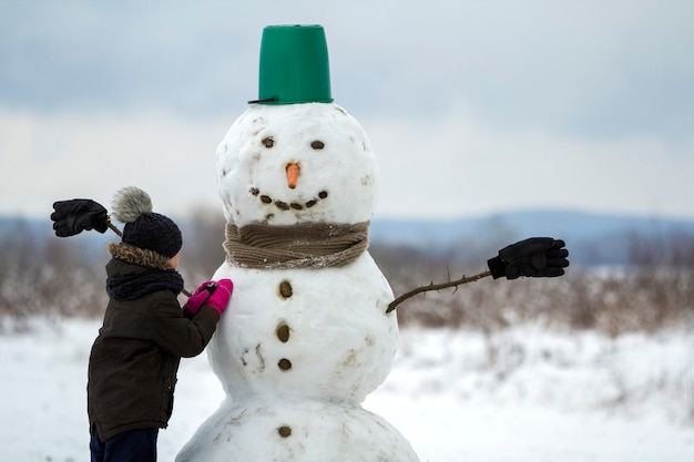 幼い子供の男の子は、空の冬の野原の風景と澄んだ青い空のコピースペースでバケツの帽子、スカーフ、手袋で笑顔の雪だるまをやり終えます。