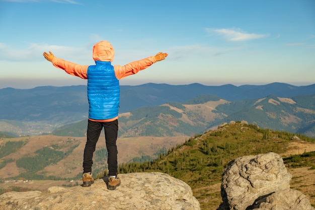 Young child boy hiker standing with raised hands in mountains enjoying view of amazing mountain landscape at sunset.