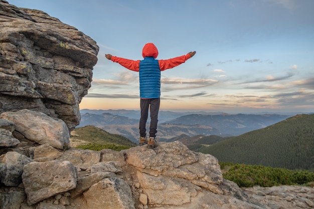 Young child boy hiker standing with raised hands in mountains enjoying view of amazing mountain landscape at sunset.