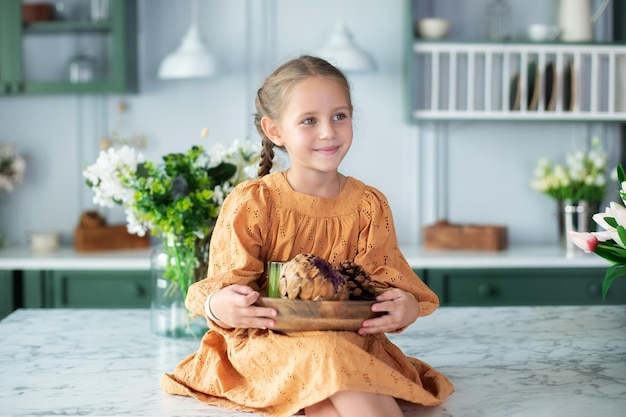 La ragazza del bambino in vestito giallo con le trecce tiene il piatto di legno con l'arredamento in cucina a casa