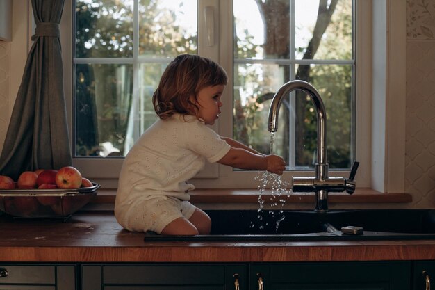 A young child is seated in a kitchen sink delighting in the water as sunlight streams