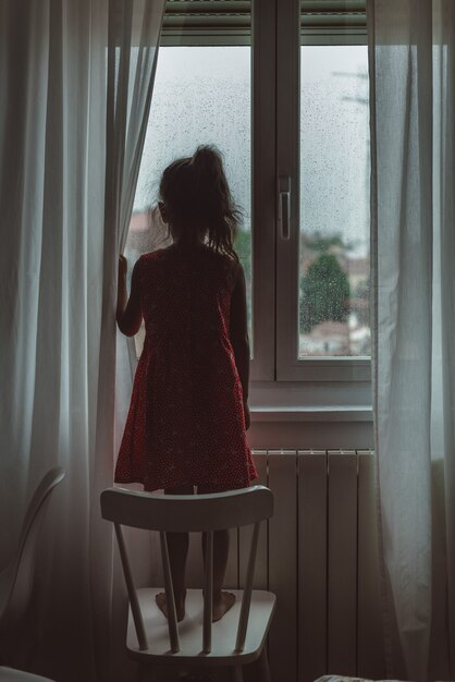 Young child in a red dress looking out of a window on a rainy day