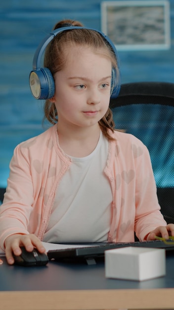 Young child wearing headphones and drawing on textbook with pencils at desk. Little girl listening to music and using computer while having notebook to draw after online school classes