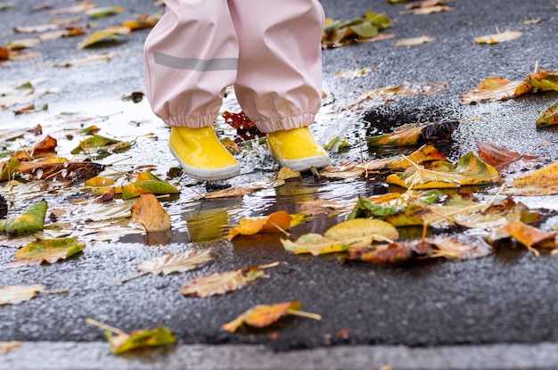 A young child with yellow boots splashes in a rain puddle with autumn leaves