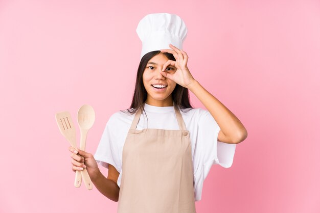 Young chinese chef woman isolated excited keeping ok gesture on eye.