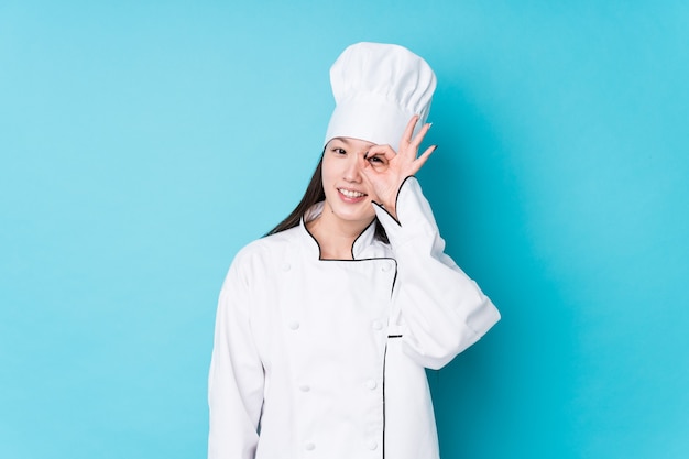 Young chinese chef woman isolated excited keeping ok gesture on eye.