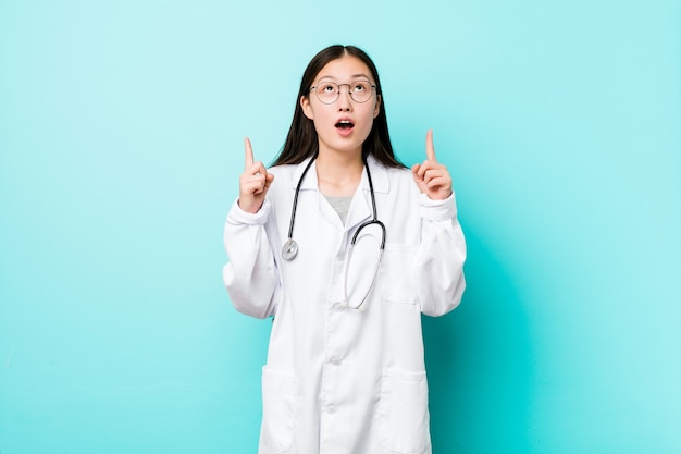 Young chinese doctor woman pointing upside with opened mouth.