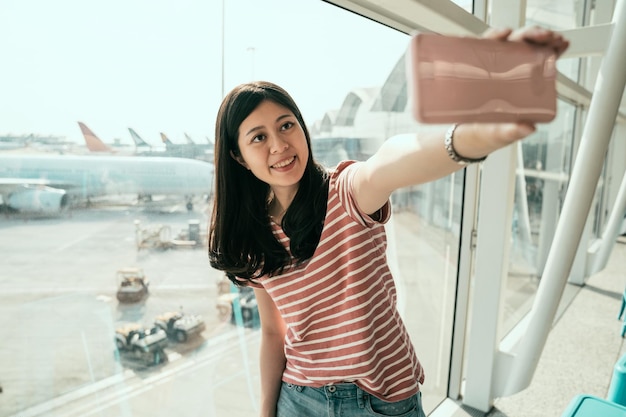 young chinese female traveler in airport lounge near windows happy smile taking selfie photo flight. happy girl tourist holding cellphone using camera app making self portrait with airplane on runway