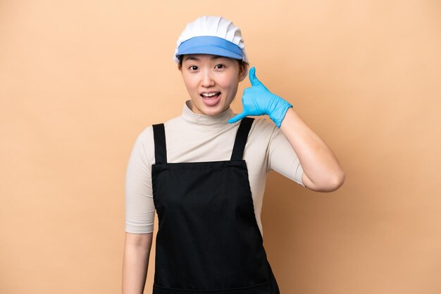 Young Chinese Fishmonger woman wearing an apron and holding a raw fish isolated on pink background making phone gesture Call me back sign