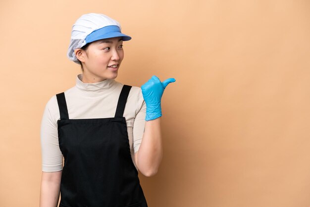 Young Chinese Fishmonger woman wearing an apron and holding a raw fish isolated on pink background pointing to the side to present a product