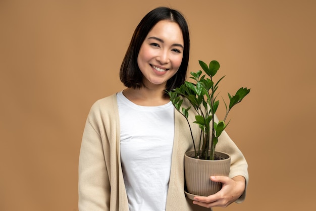 ベージュの背景に分離された喜びの笑顔で緑の植物を保持している若い中国の庭師の女性、彼女は笑顔と陽気な感じ