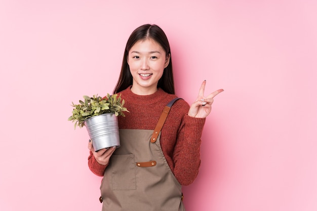 Young chinese gardener woman holding a plant isolated joyful and carefree showing a peace symbol with fingers.