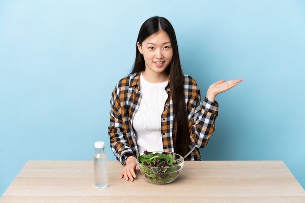 Молодая китаянка ест салат, держа на ладони воображаемое пространство, чтобы вставить рекламу
