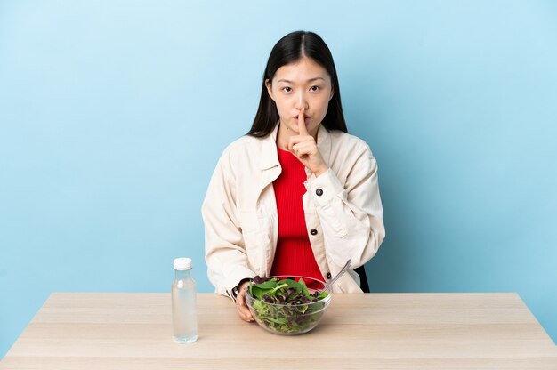 Young Chinese girl eating a salad showing a sign of silence gesture putting finger in mouth