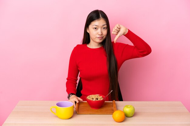 부정적인 표현으로 아래로 엄지 손가락을 보여주는 테이블에서 아침을 먹고 젊은 중국 소녀